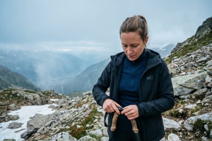 Randonneuse portant une veste technique de marque française sur un sentier alpin rocheux avec vue sur les sommets enneigés